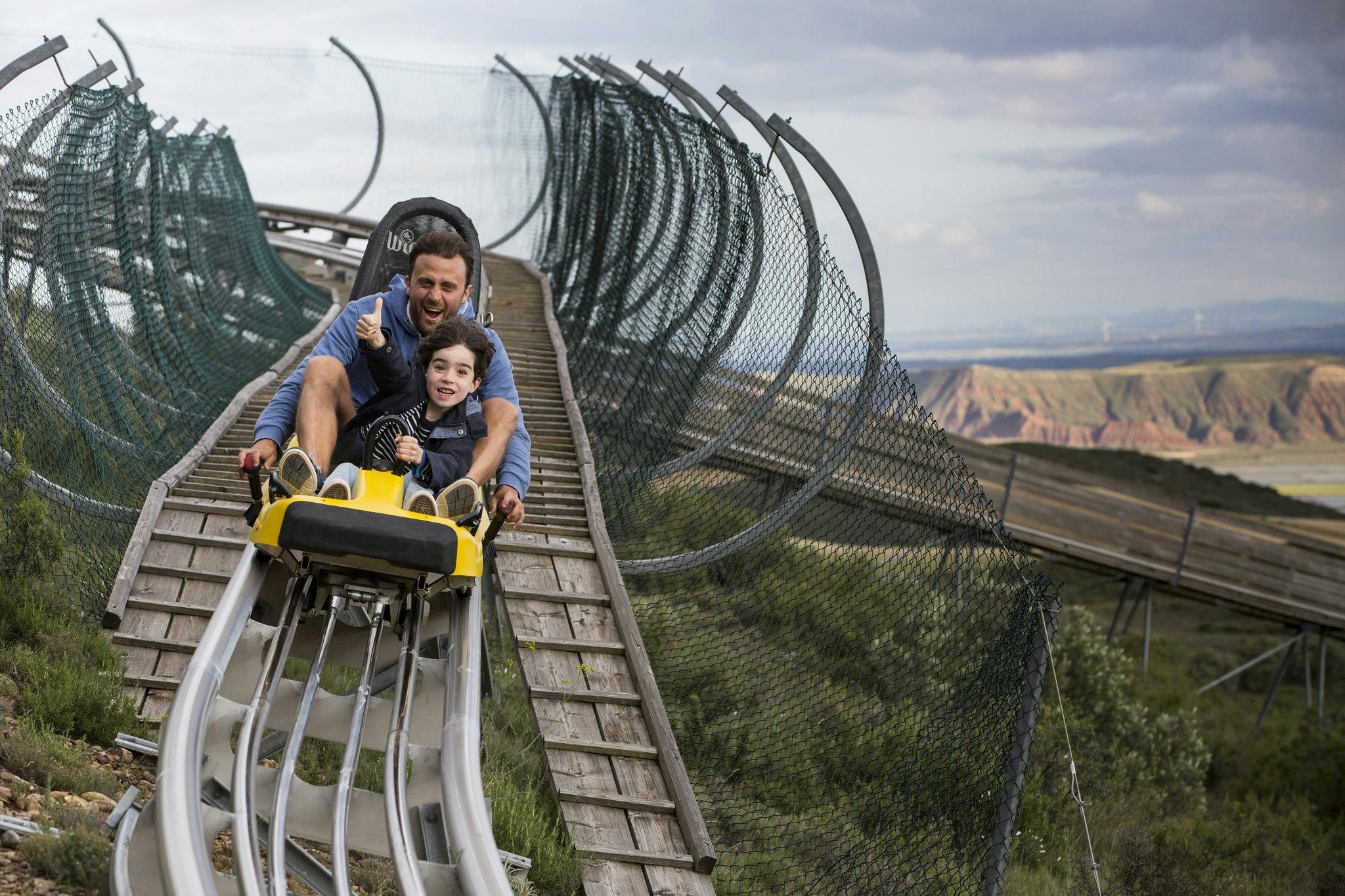 Muž a dítě jedou na žluté horské dráze po dřevěné dráze obklopené síťovinou s hornatým terénem v pozadí.