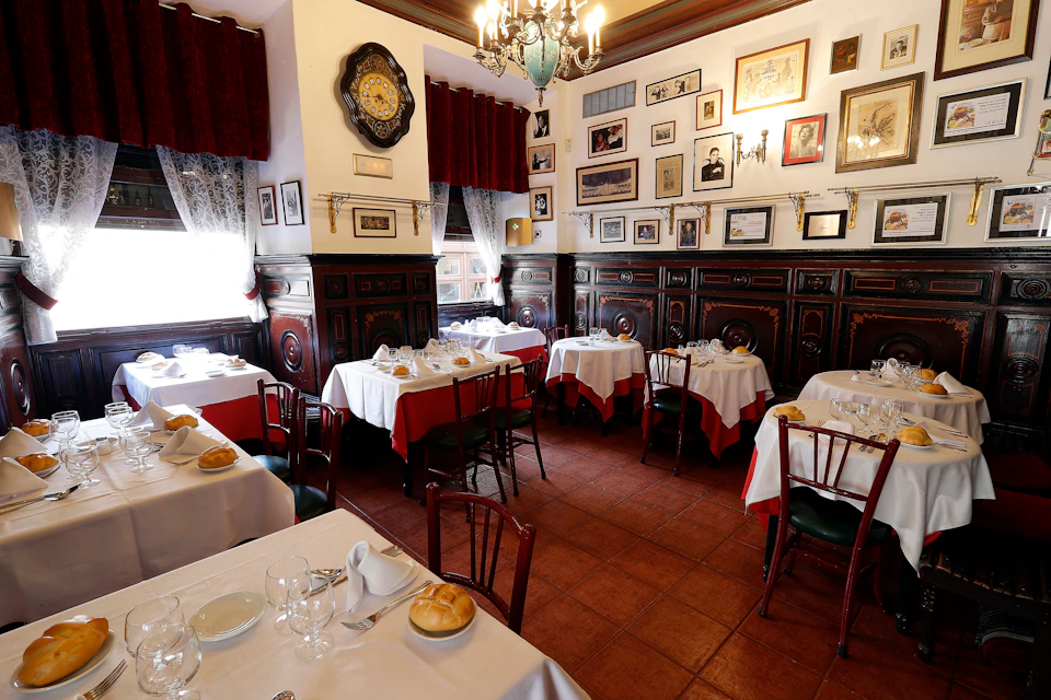 A cozy restaurant with framed pictures on the walls, white tablecloths with red accents, set tables, and wooden chairs.