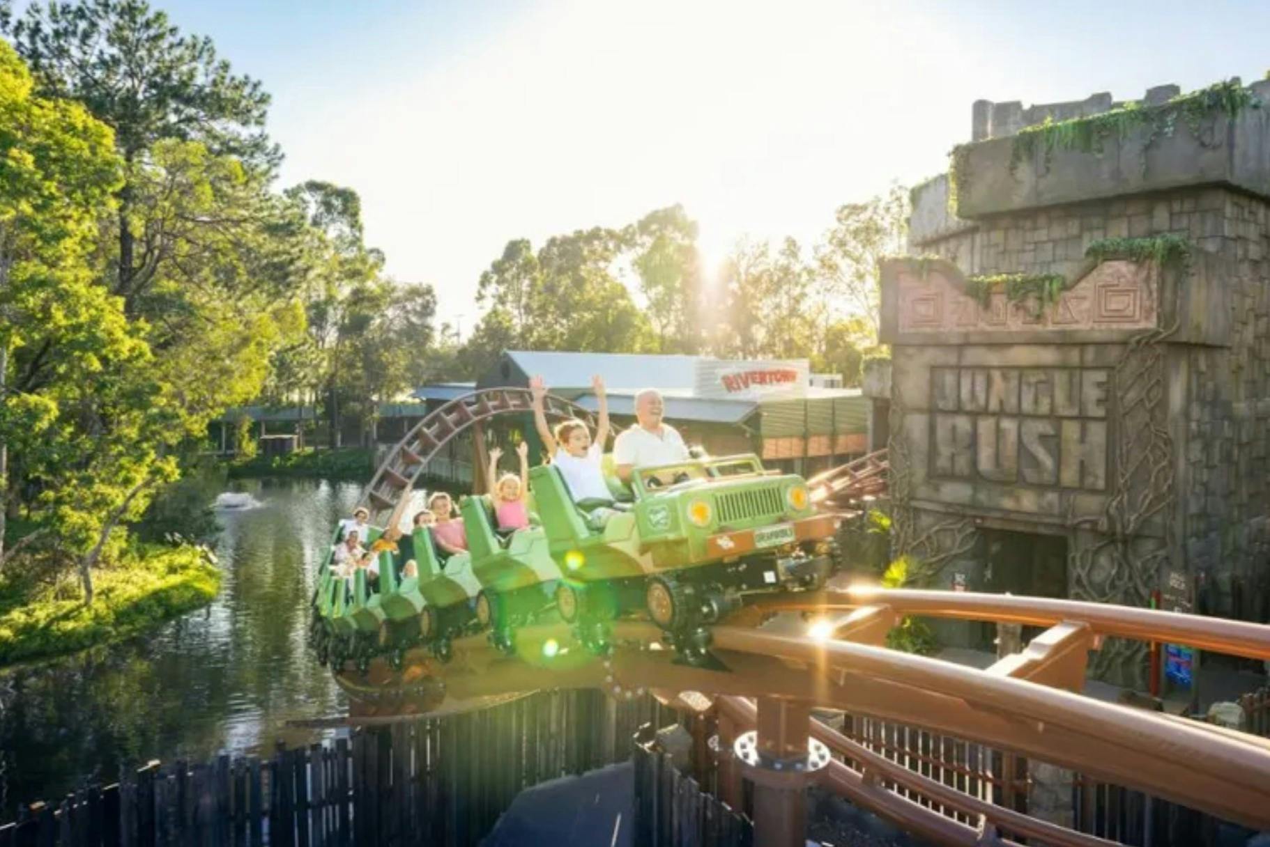 A roller coaster shaped like green jeeps travels along a track with passengers raising their arms, surrounded by greenery.