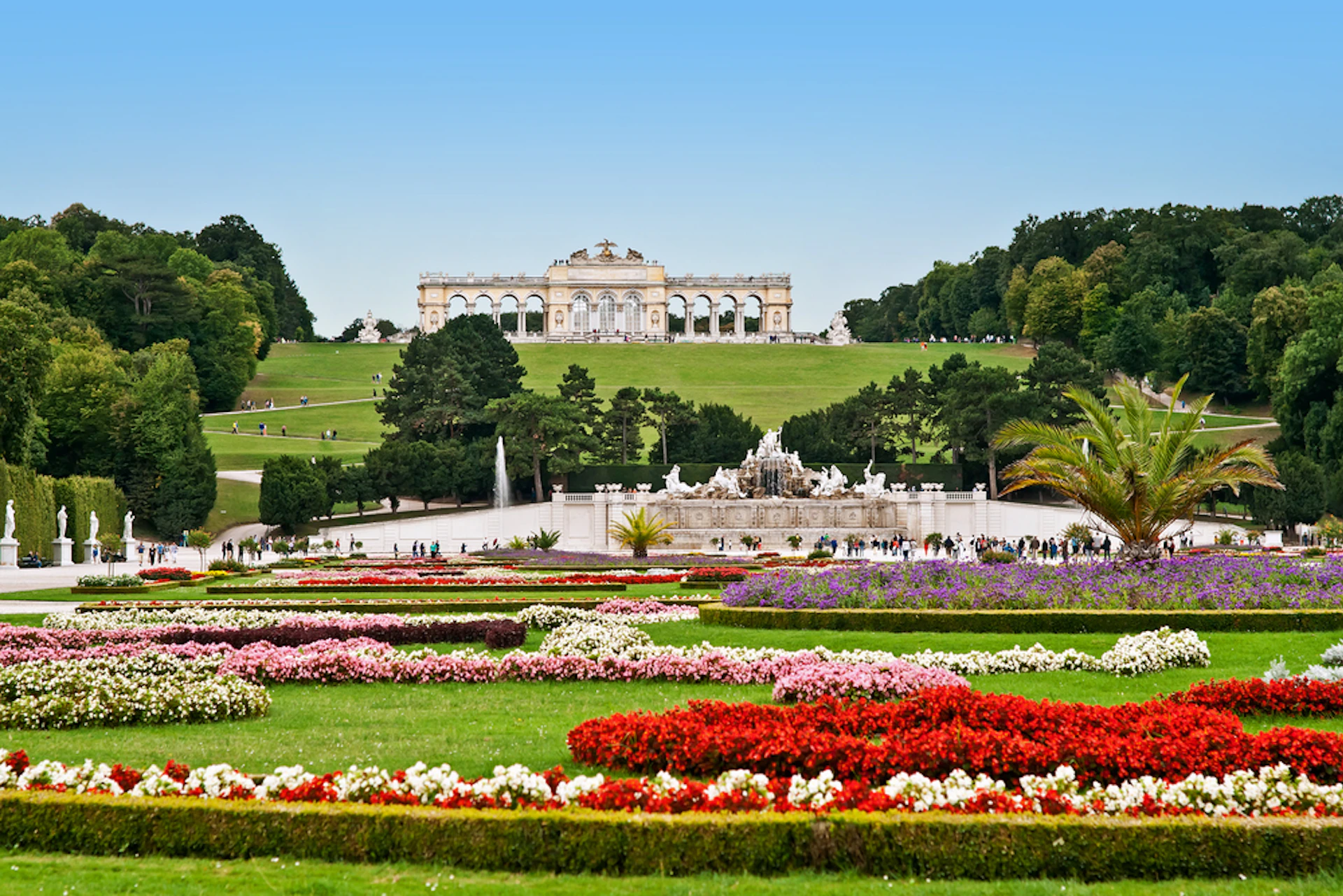 Colorful flowerbeds, fountain, and landscaped greenery leading to a grand, ornate building on a hill under a clear blue sky.