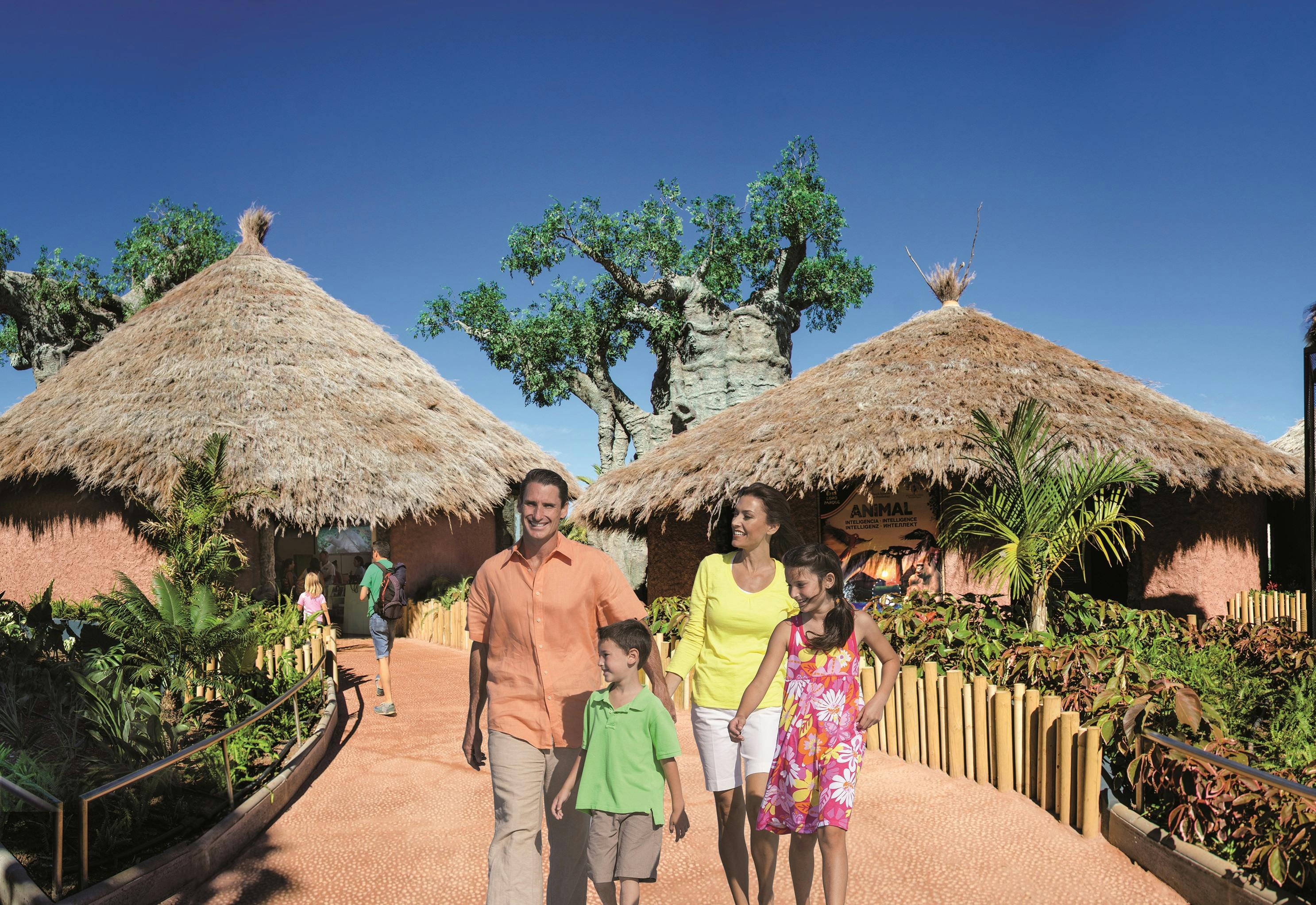4인 가족이 맑고 푸른 하늘 아래 열대 식물과 초가 지붕 오두막이 늘어선 길을 걷고 있습니다.
