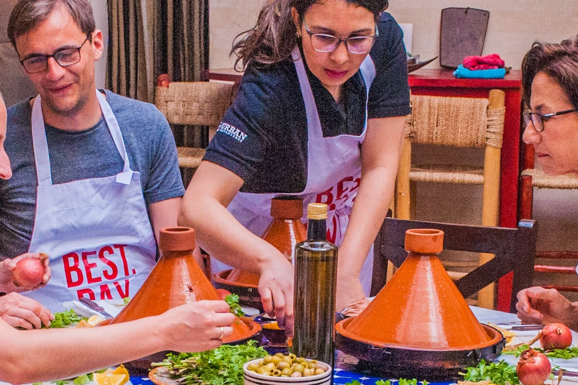 People wearing aprons preparing ingredients around a table with vegetables and tagines in a brightly lit kitchen.