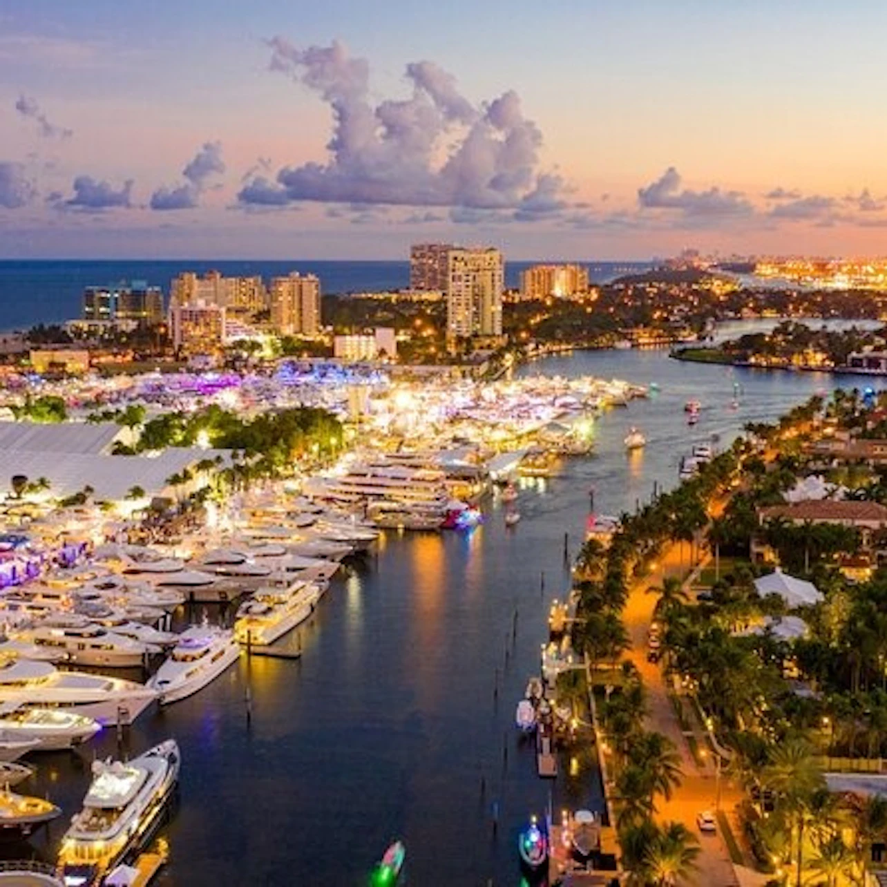 A vibrant marina at sunset with numerous yachts, palm trees, and city lights in the background.
