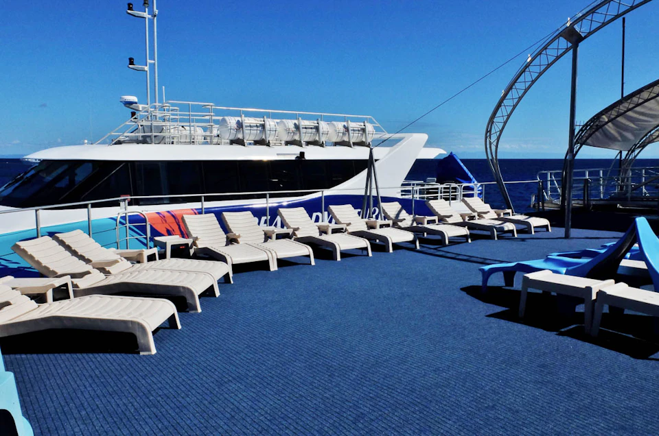 A row of empty lounge chairs on a blue deck facing a docked, white yacht under a clear blue sky.