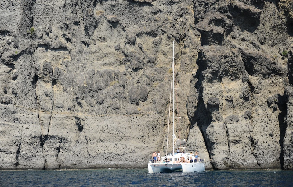 A catamaran with people on board is anchored near a large rocky cliff face, with calm water in the foreground.