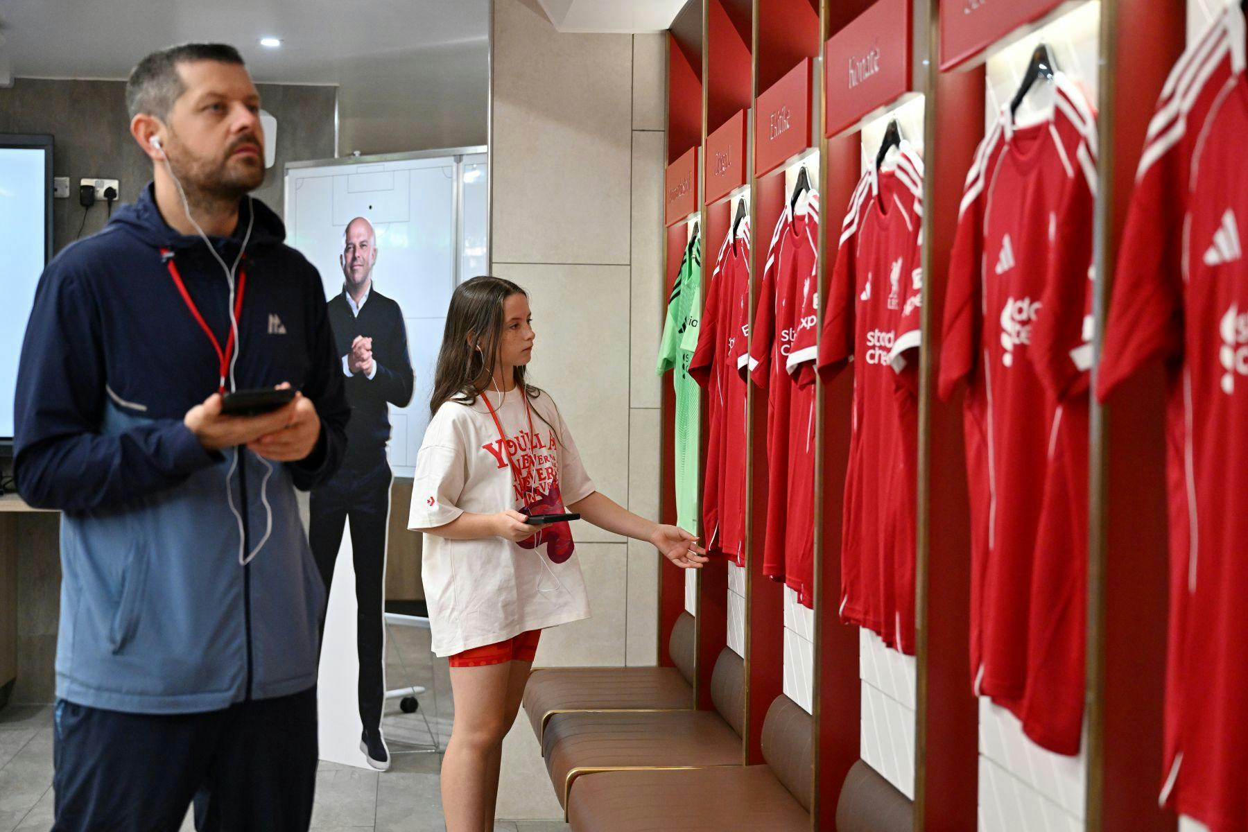 라커룸에서 축구 유니폼을 바라보며 이어폰과 휴대폰을 든 남성과 스포츠웨어를 입은 소녀. 백그라운드에서 골판지 컷아웃.