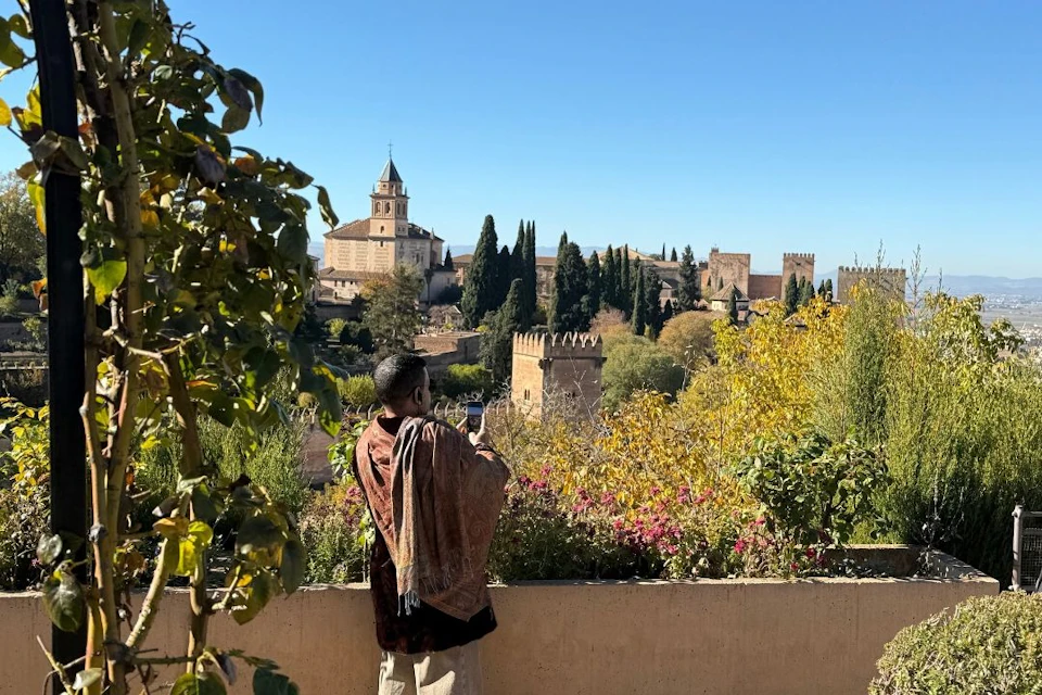A person taking a photo of a scenic cityscape featuring historic buildings, trees, and a clear blue sky.