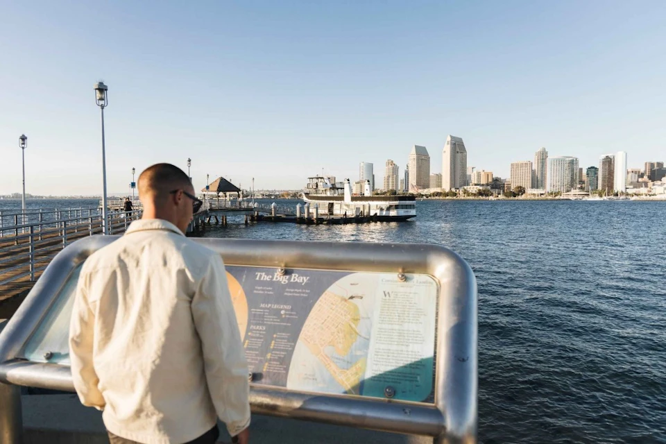 A person reads an informational sign at a waterfront, with a city skyline and a pier visible across the water.