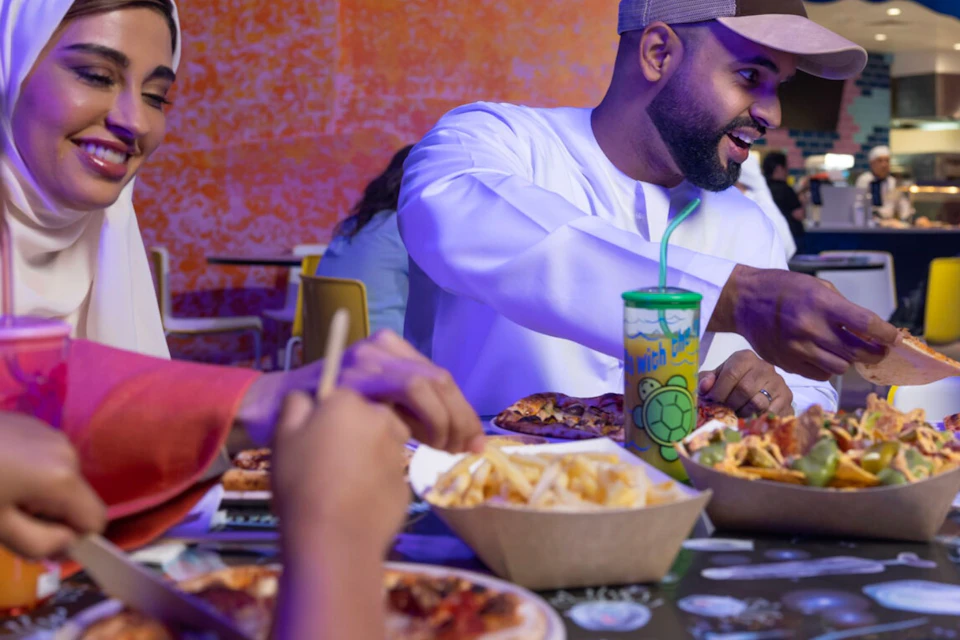 A group of people enjoying a meal at a restaurant under a sign reading 'Bitsy Bites,' with various food items on the table.
