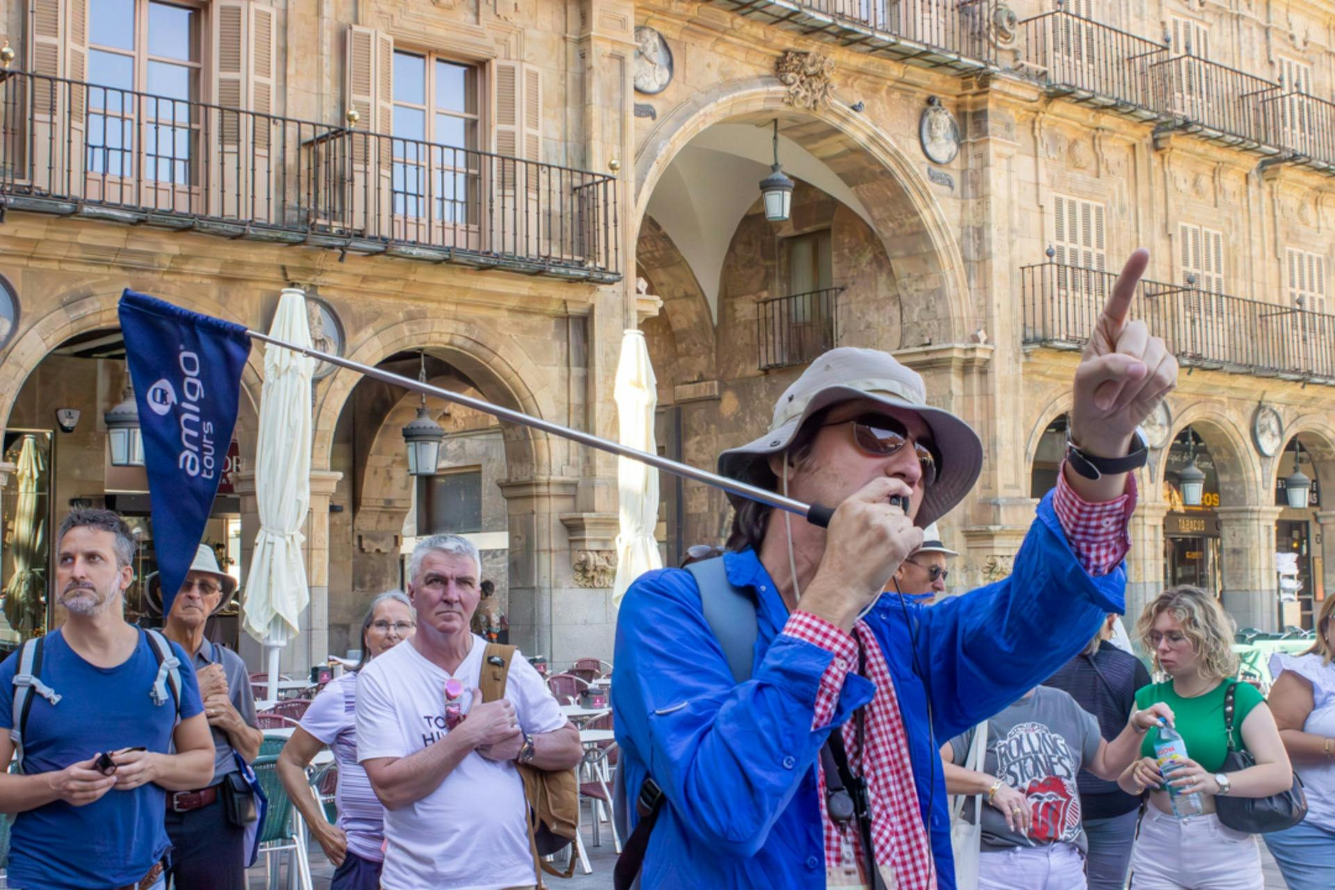 Μια ομάδα ανθρώπων σε μια ξενάγηση σε μια ιστορική πλατεία, με έναν οδηγό που κρατά μια σημαία και φοράει ένα καπέλο για τον ήλιο σε πρώτο πλάνο.