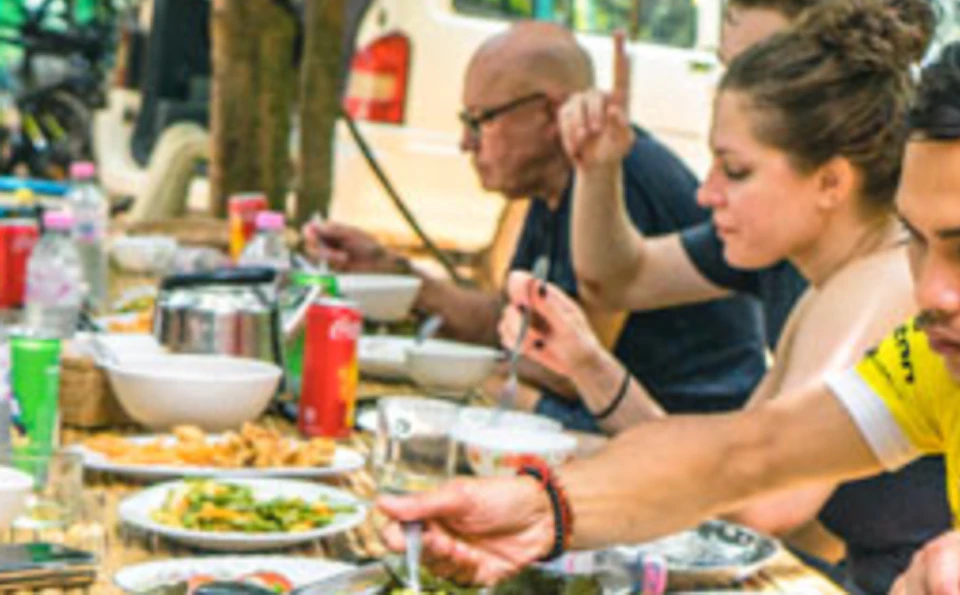 Four people are seated at a wooden table outdoors, eating a meal with various dishes in a shaded setting surrounded by trees.