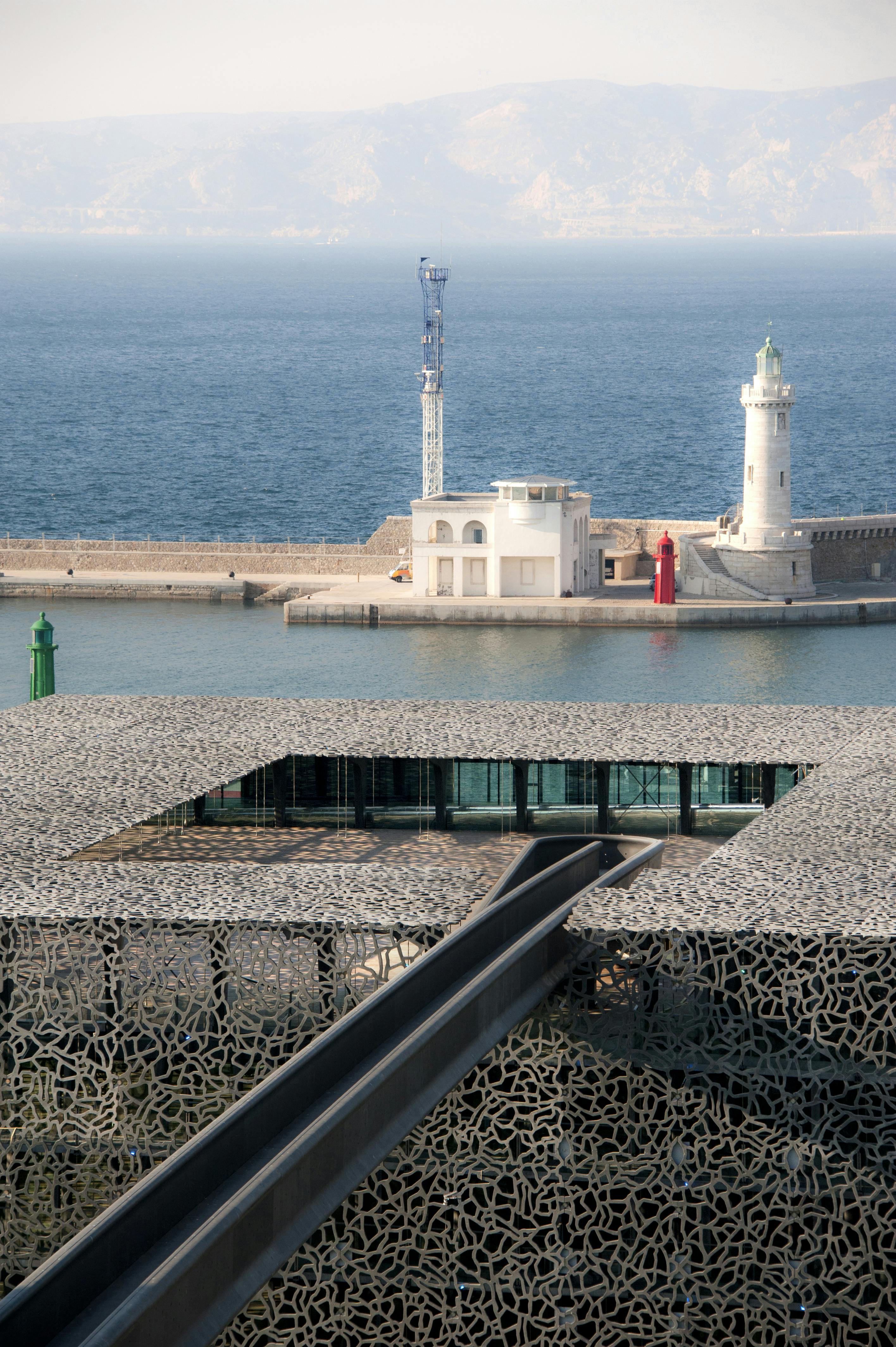 Nowoczesny budynek z wzorzystym dachem z widokiem na wodę, z latarnią morską i małym białym budynkiem na falochronie w tle.