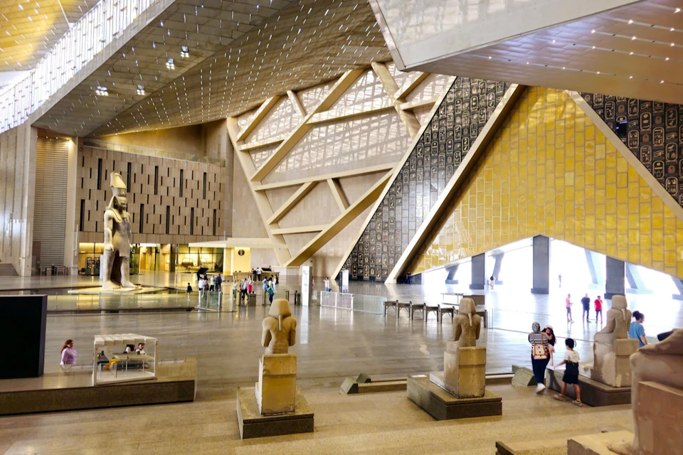 A modern museum interior with large geometric design elements, statues, and visitors walking through the space.