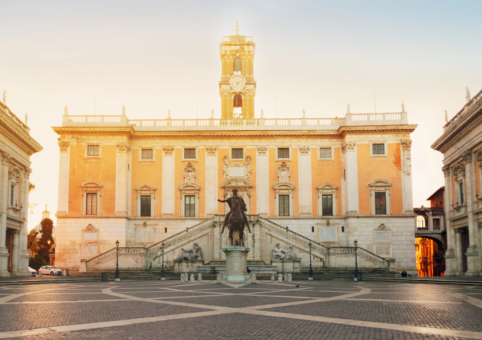 A historic building with a central clock tower, grand staircase, and equestrian statue in the foreground, under a clear sky.