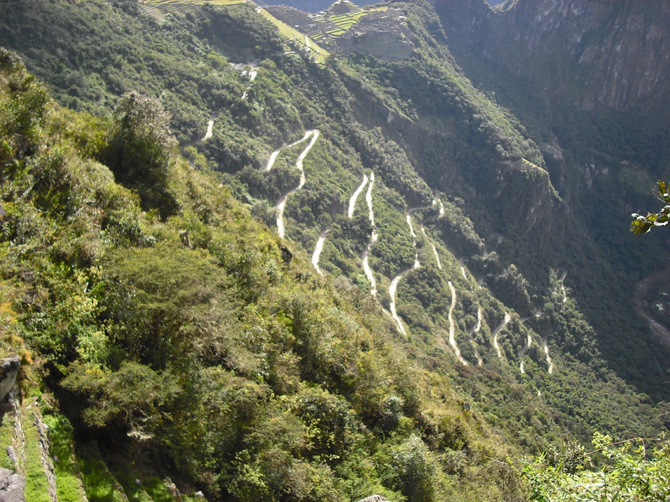 A winding, zigzagging road ascends a steep, lush green mountainside in a mountainous landscape.
