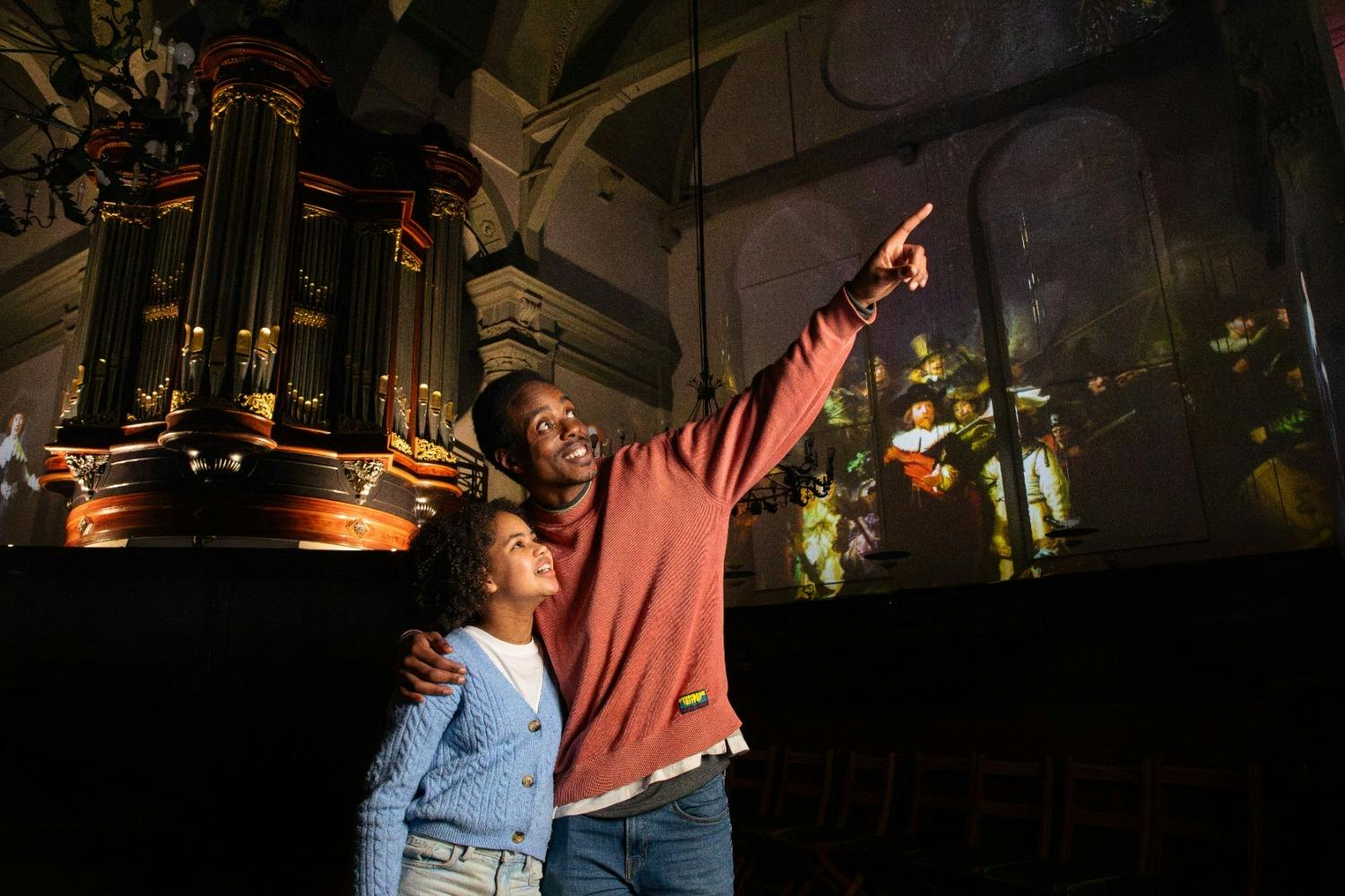 Ένας άνδρας και ένα παιδί στέκονται μπροστά από ένα μεγάλο εκκλησιαστικό όργανο, χαμογελώντας και δείχνοντας προς τις προβαλλόμενες εικόνες σε έναν τοίχο.