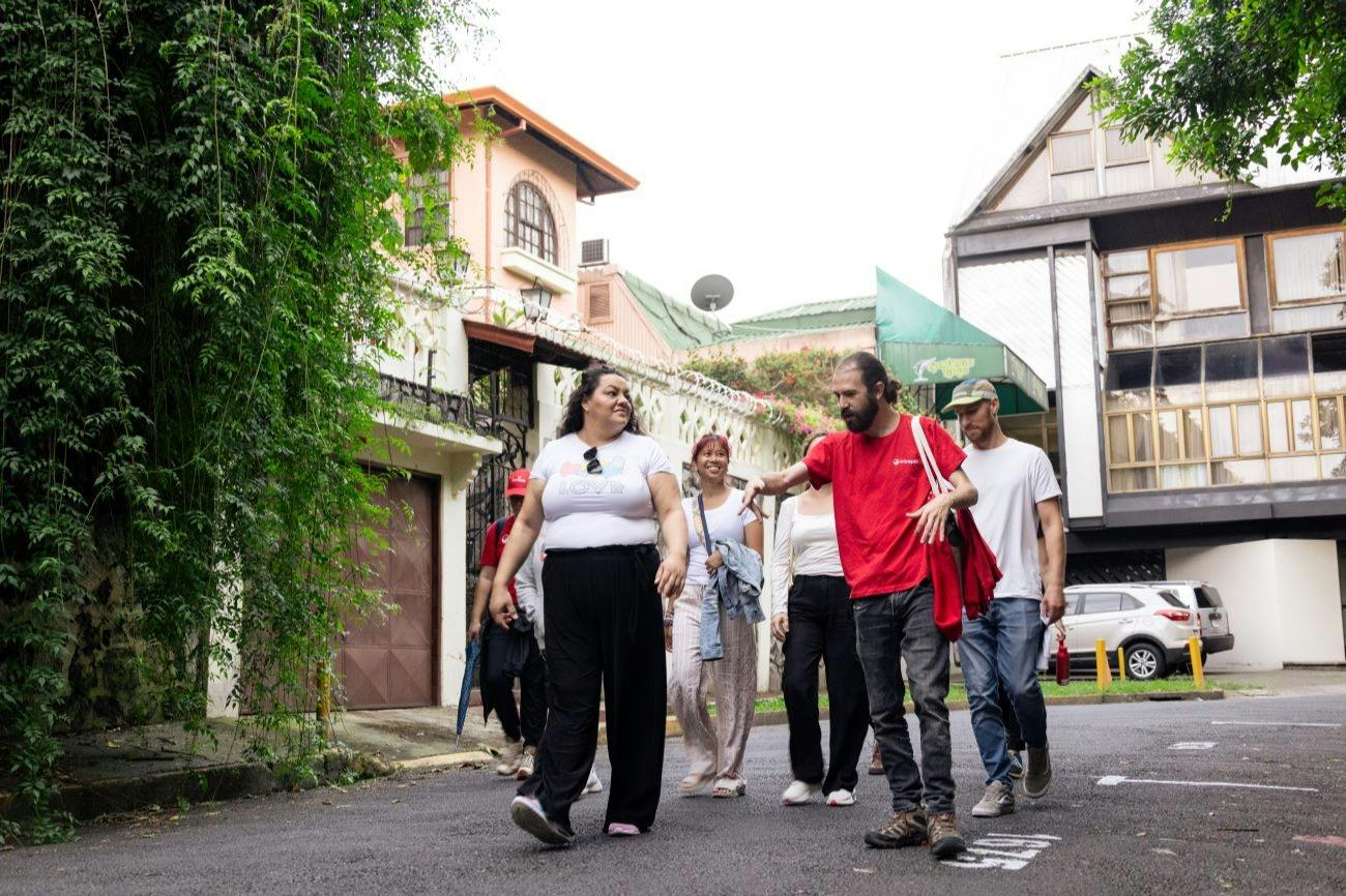 A group of people walking on a paved street near houses, with greenery and a bright sky in the background.
