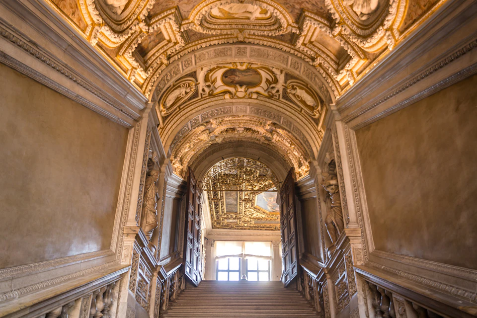 A grand staircase with ornate, gilded ceiling and open double doors leading to a sunlit window at the top.