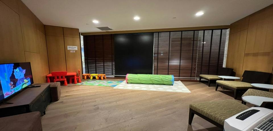 A play area with a green play tunnel, colorful chairs, and a play mat on a wooden floor near window blinds.