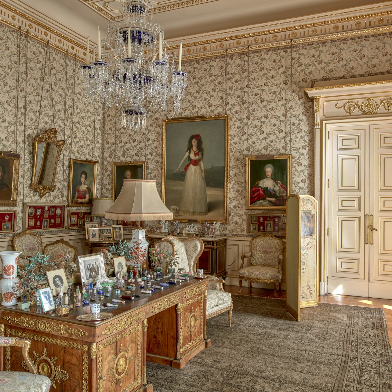 Utsmyckat rum med blommiga tapeter, ljuskrona, stora målningar, mönstrade stolar och ett träbord med inramade foton och små flaskor.