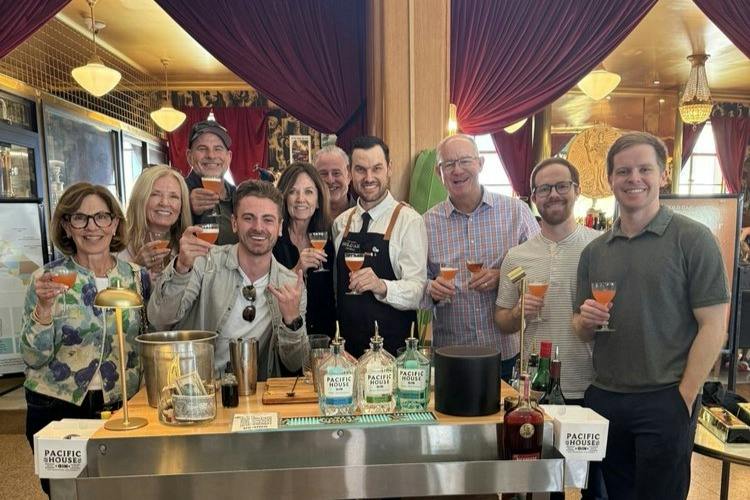 A group of people smiling and holding cocktails around a bar counter with bottles and mixers, in a warmly lit room.