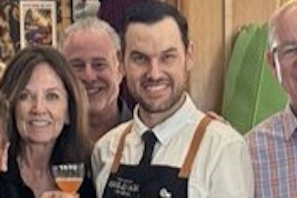 A group of people smiling and holding cocktails around a bar counter with bottles and mixers, in a warmly lit room.