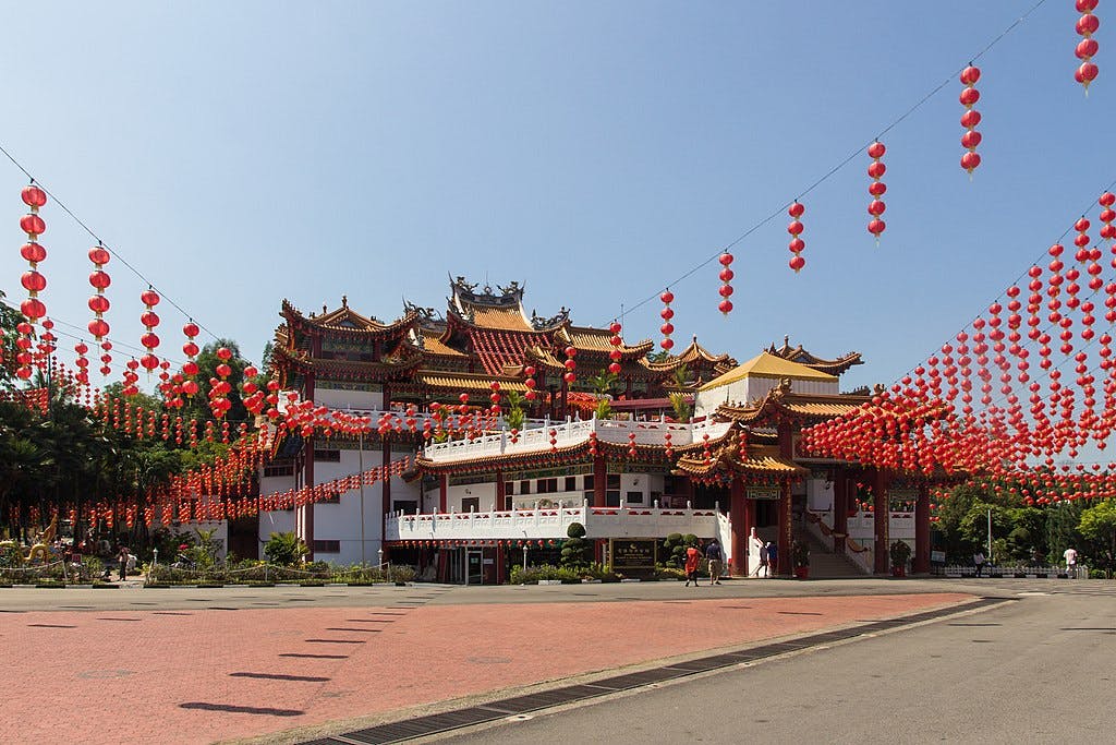 A temple with intricate roof designs adorned with numerous red lanterns under a clear blue sky. Few people are visible.