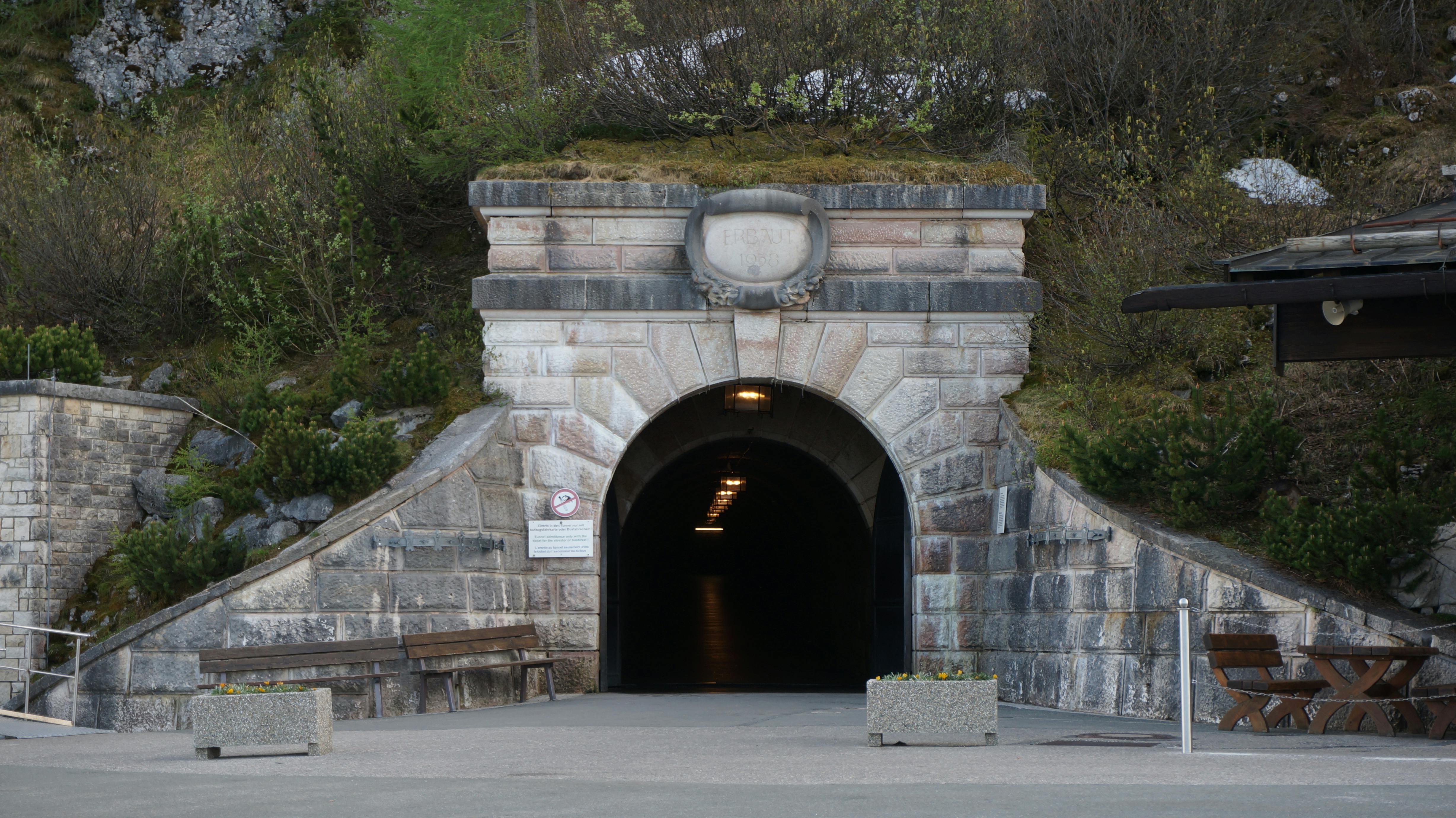 Vstup do kamenného tunelu s klenutým otvorem, lemovaný kamennými zdmi a zelení, vedoucí do temné chodby.