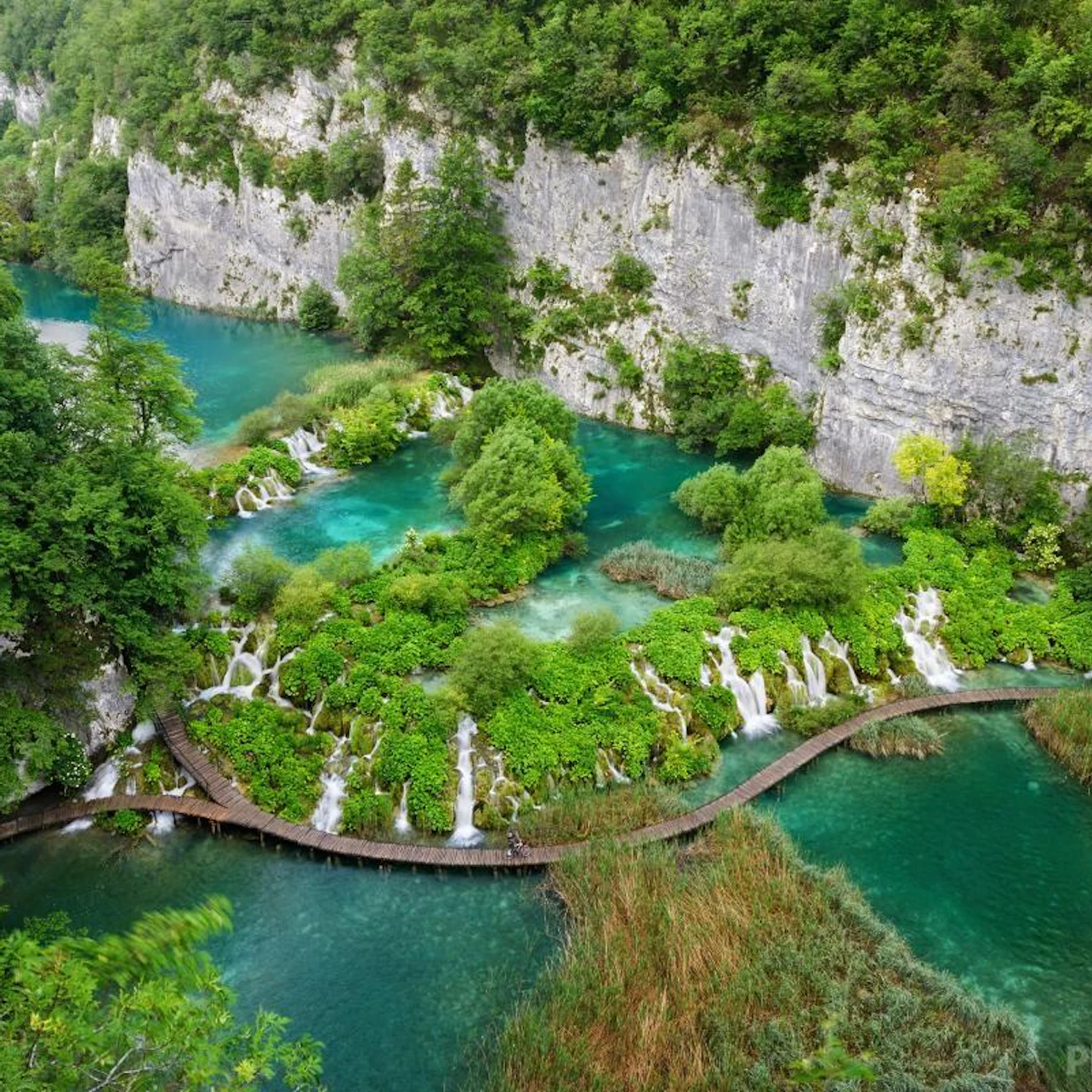 A lush landscape with turquoise lakes, waterfalls, and a wooden path winding through greenery and rocky cliffs.