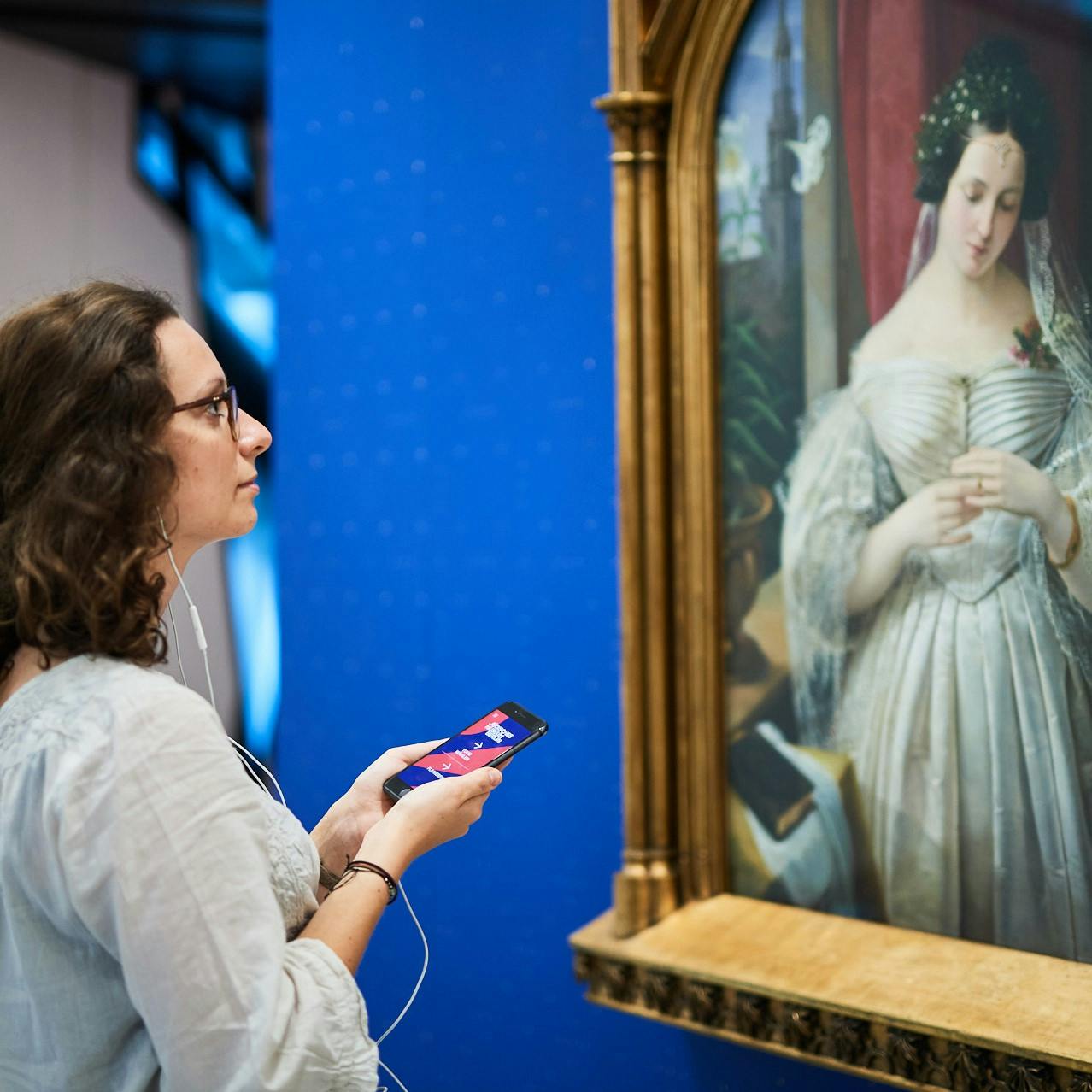 Osoba s brýlemi, držící chytrý telefon a nosící sluchátka, dívající se na klasický obraz ženy v bílých šatech.