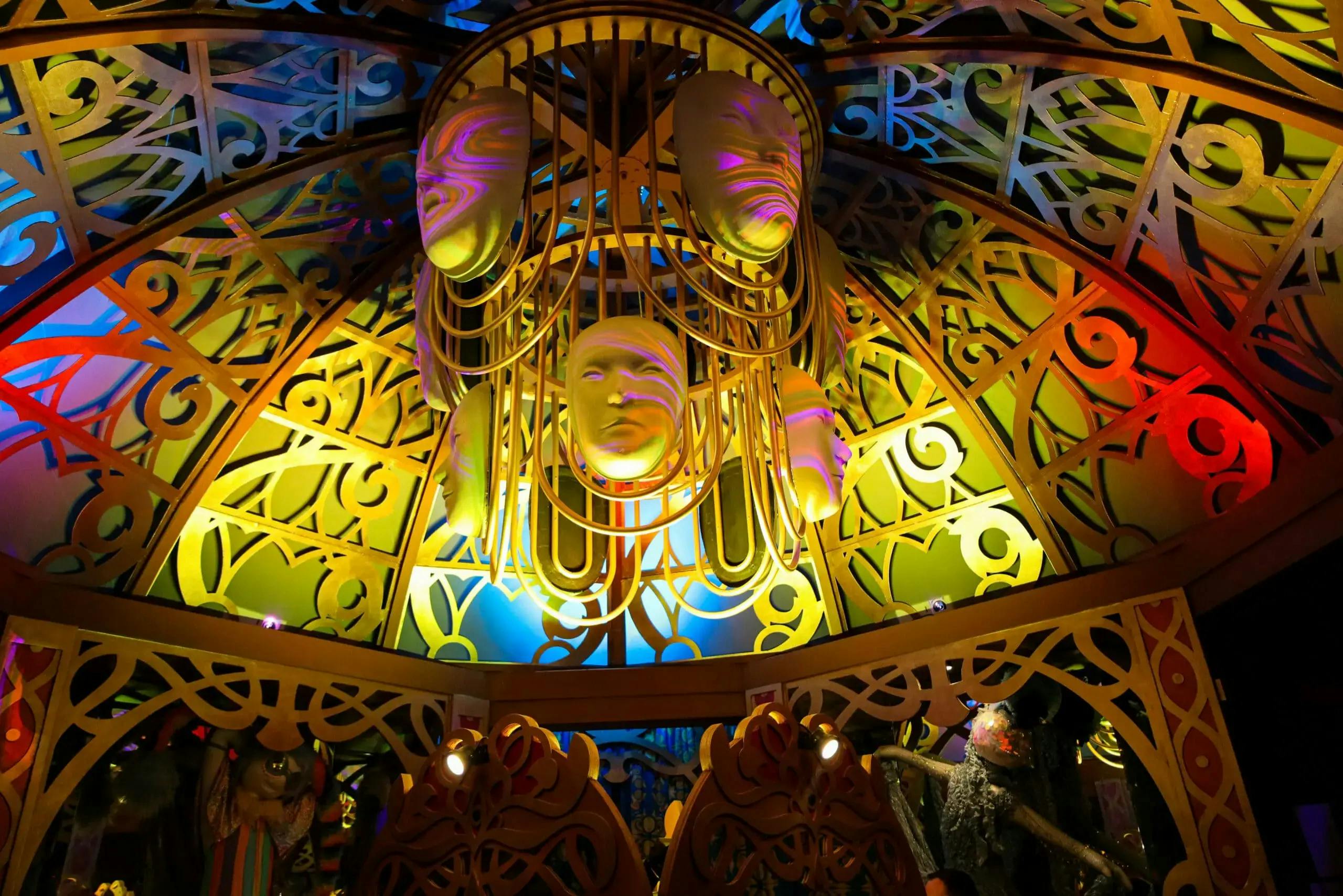 A ceiling adorned with yellow theatrical masks and intricate patterns, illuminated by colorful lights, creating a whimsical atmosphere.
