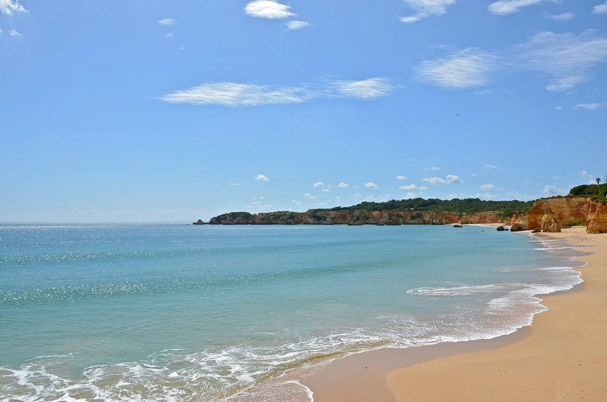 A tranquil sandy beach with gentle waves, clear blue water, and a distant rocky coastline under a bright blue sky.