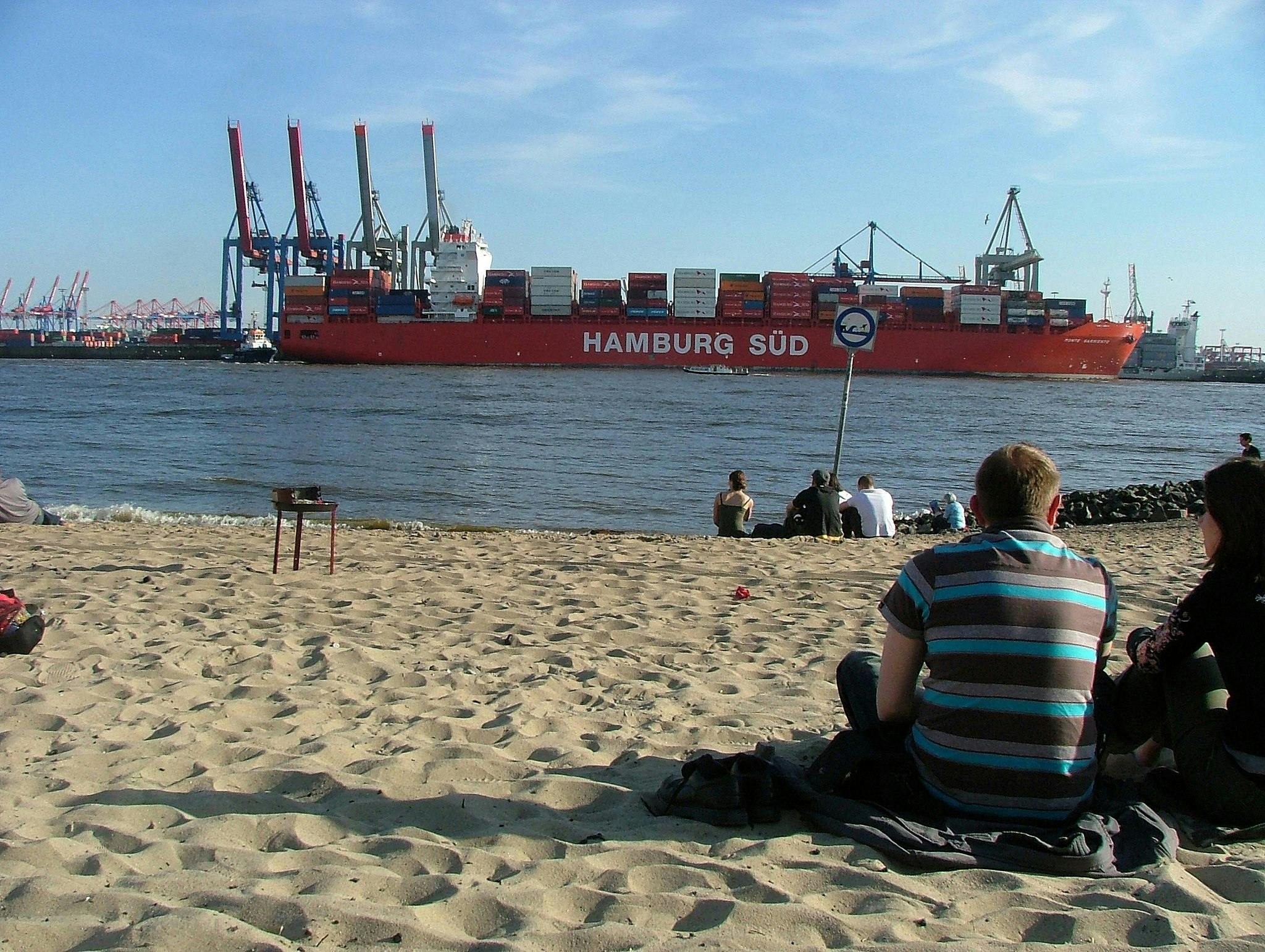 Persone sedute su una spiaggia sabbiosa che guardano una grande nave portacontainer rossa con la scritta "Hamburg Süd" passare su un fiume sotto un cielo limpido.