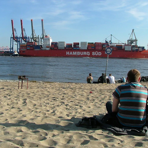 Menschen sitzen an einem Sandstrand und beobachten, wie ein großes rotes Containerschiff mit der Aufschrift "Hamburg Süd" auf einem Fluss unter einem klaren Himmel vorbeifährt.