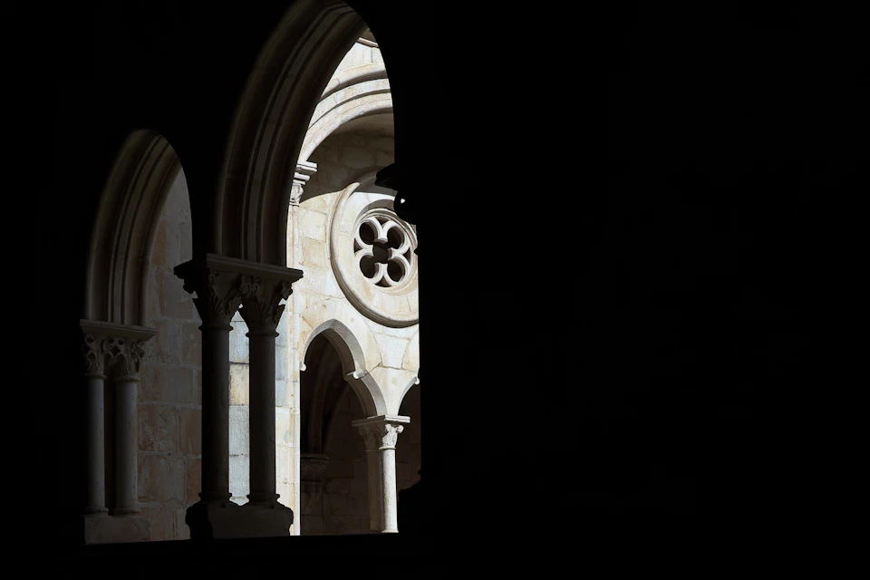 A view of a stone archway with a decorative circular window and columns, partially lit, with dark shadows in the foreground.