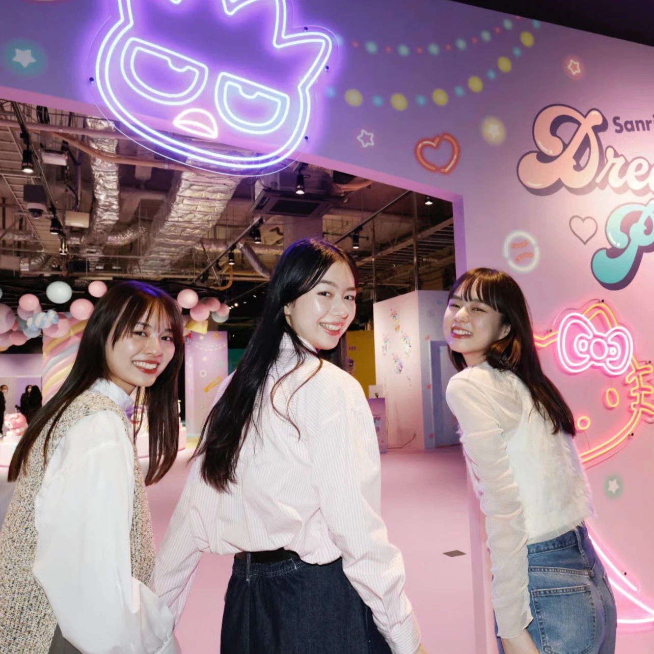 Three women smiling and posing in a colorful indoor exhibition with neon signs and playful decorations.