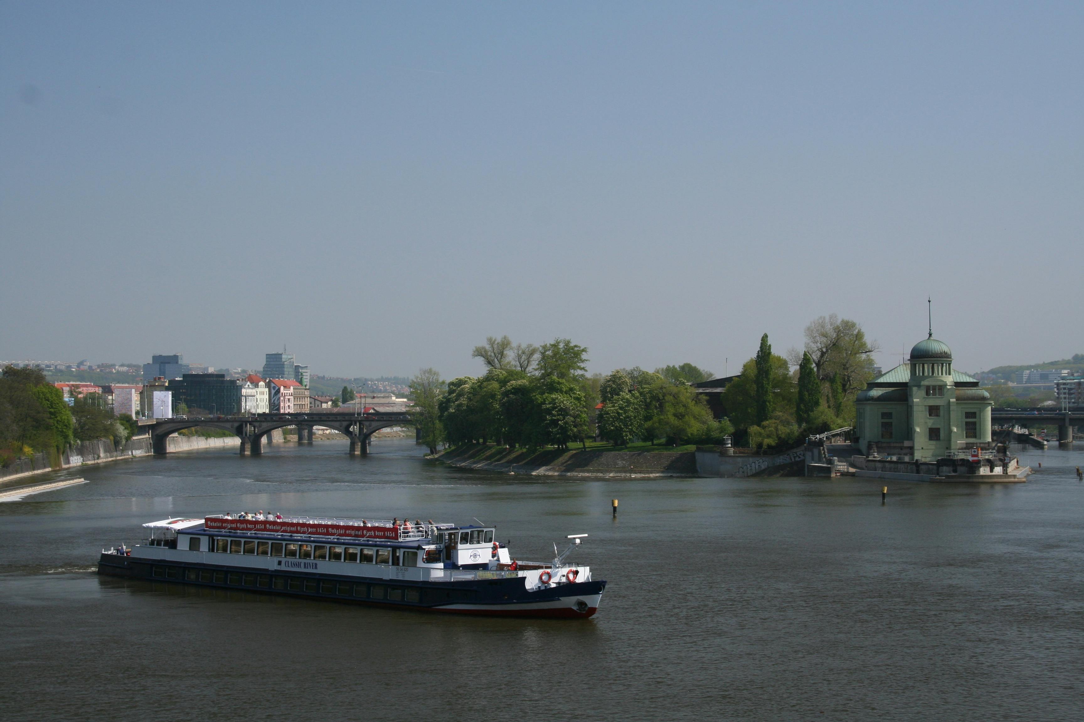 一艘河船在平静的河流上航行，在晴朗的蓝天下，后面有一座桥和绿色的树木。