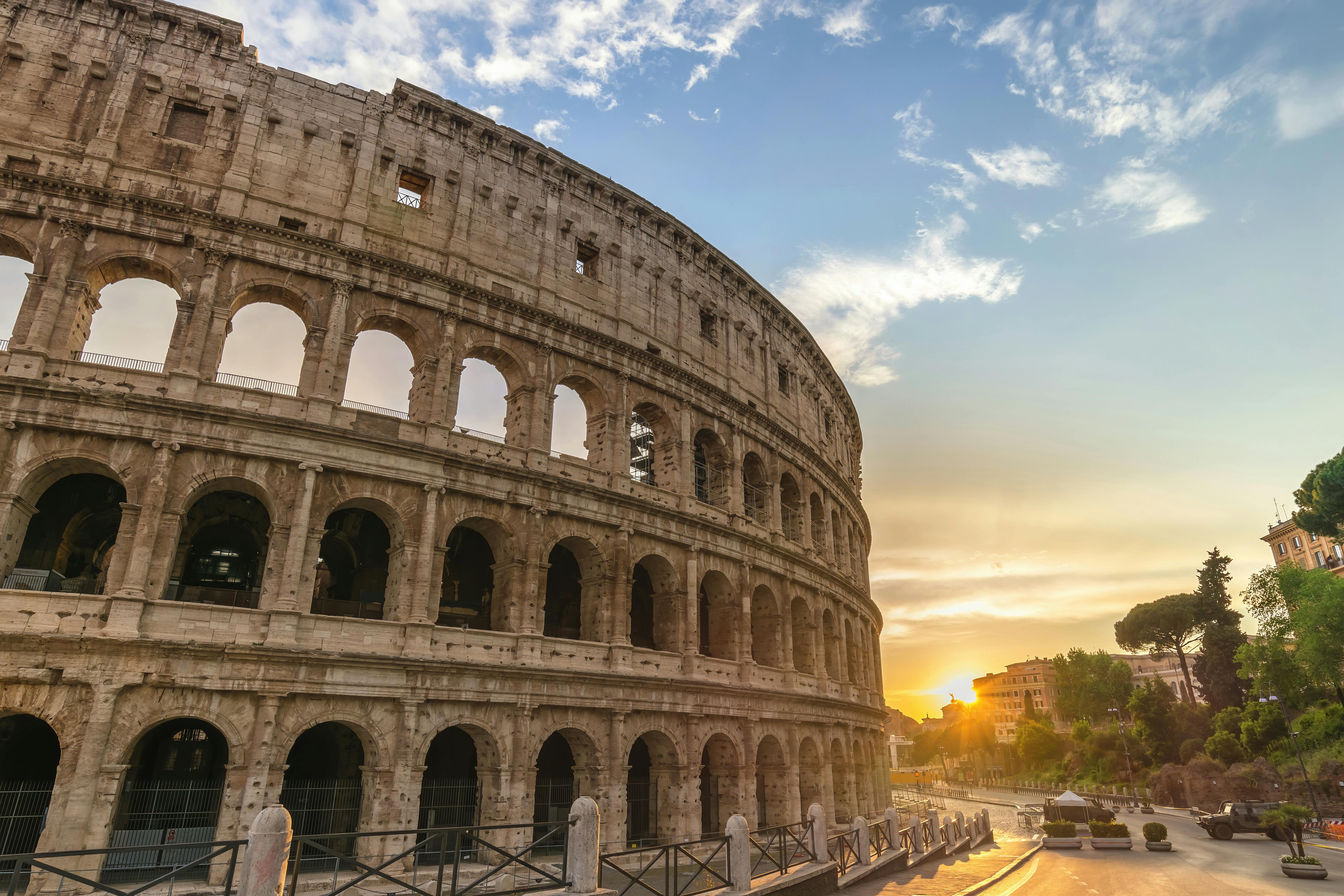 The Colosseum with its arches and stone structure is illuminated by a setting sun under a partly cloudy sky. A fence is in the foreground.