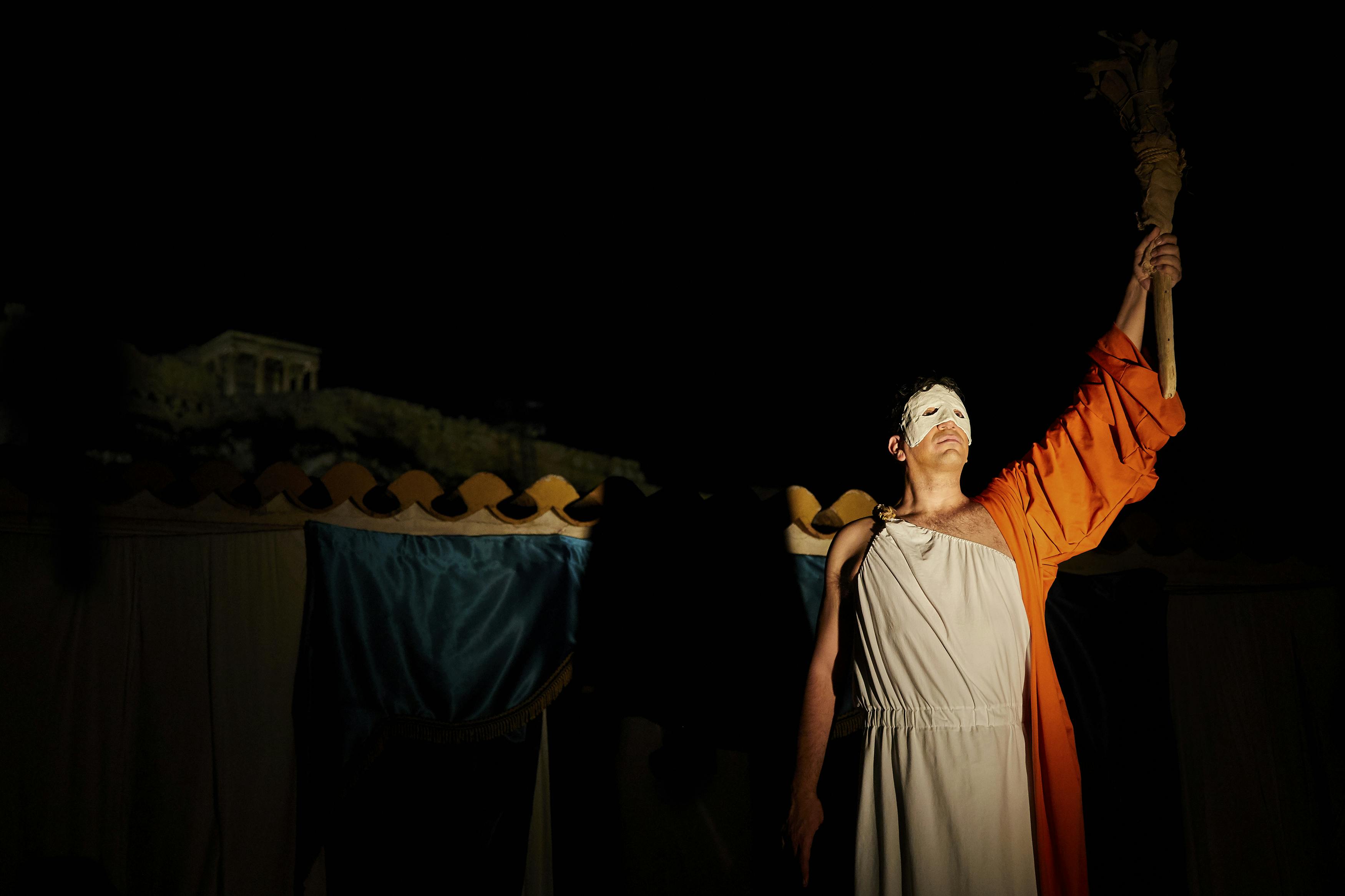 一个穿着白色长袍和橙色斗篷的人，戴着白色面具，在深色背景下高举火炬。