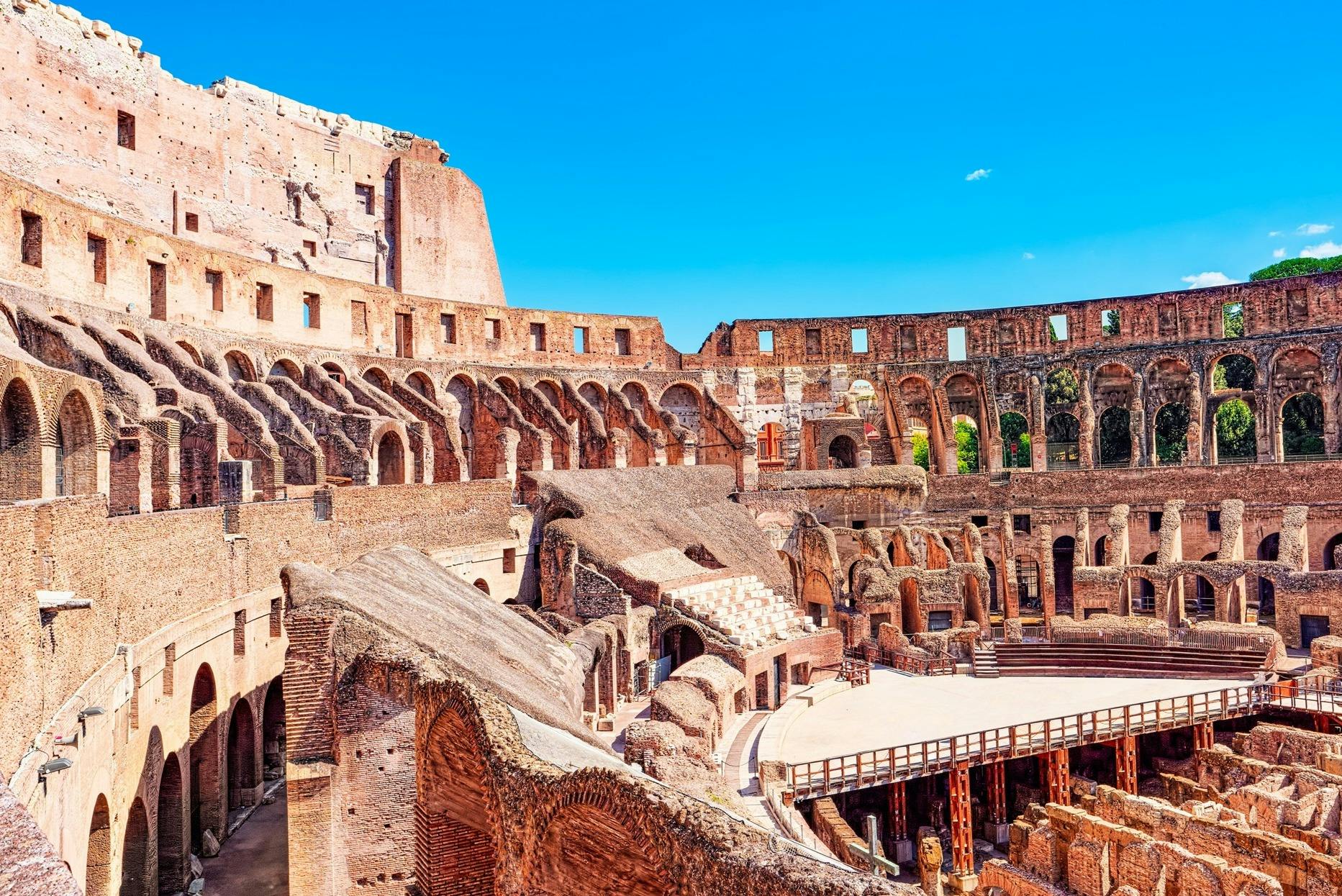 Indvendig udsigt over Colosseum i Rom, der viser gamle stenbuer, siddepladser og en del af arenaen under en blå himmel.