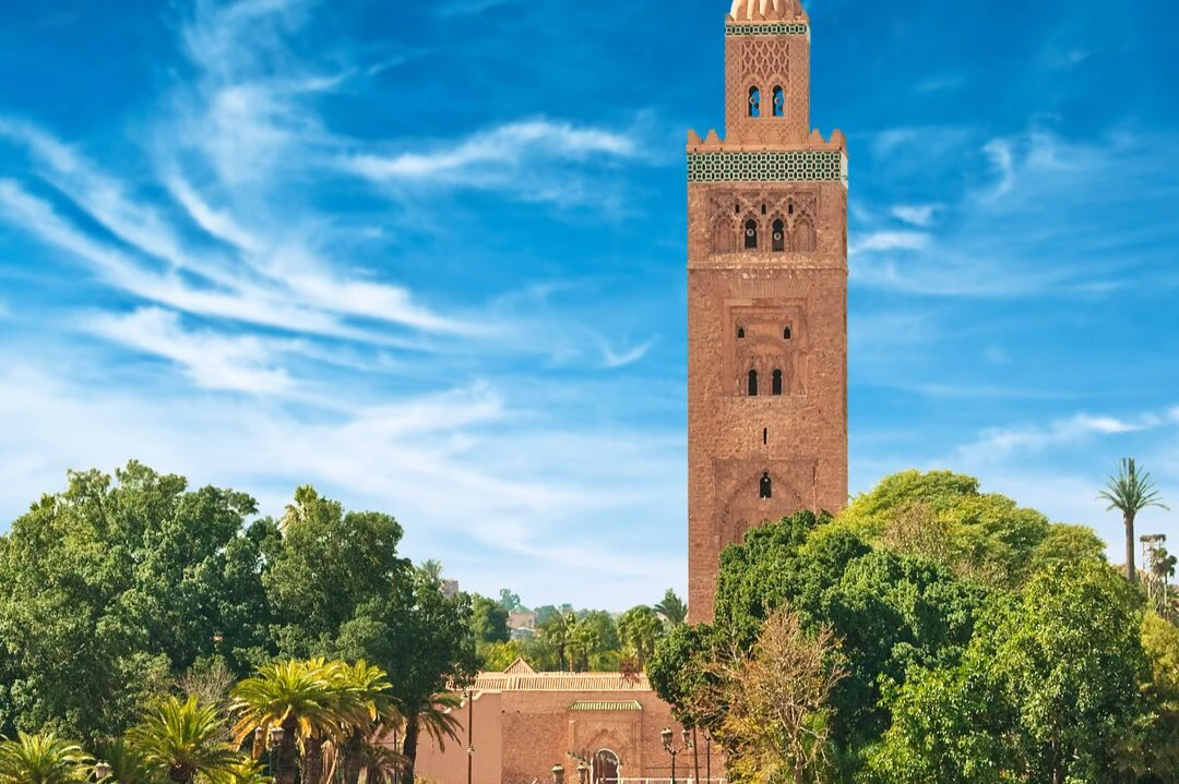 Tall brick minaret tower, surrounded by lush greenery, under a bright blue sky with scattered white clouds.