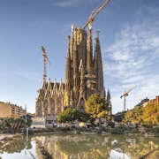 L'immagine mostra una grande cattedrale incompiuta con gru, vista da un parco con alberi rigogliosi e uno stagno riflettente.