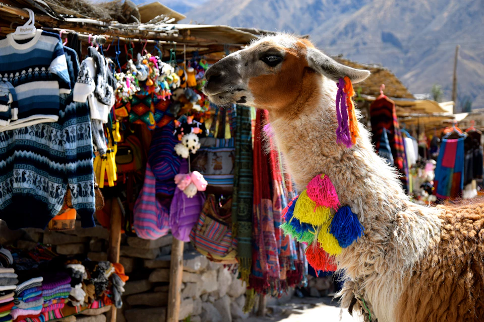 A llama with colorful tassels stands near a market stall displaying vibrant textiles and handicrafts.