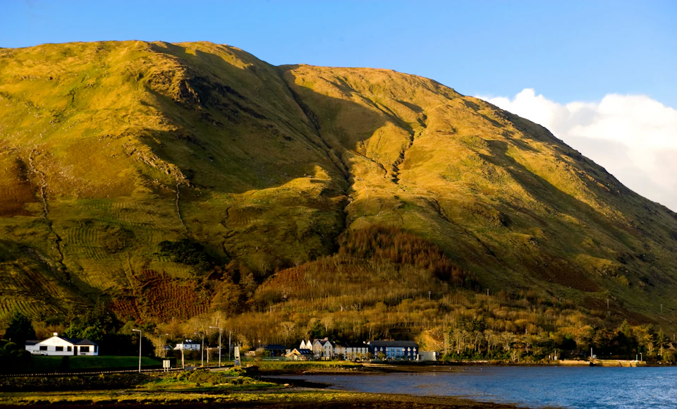 A village with several houses at the base of a large, sunlit mountain with a blue sky above. A body of water in the foreground.