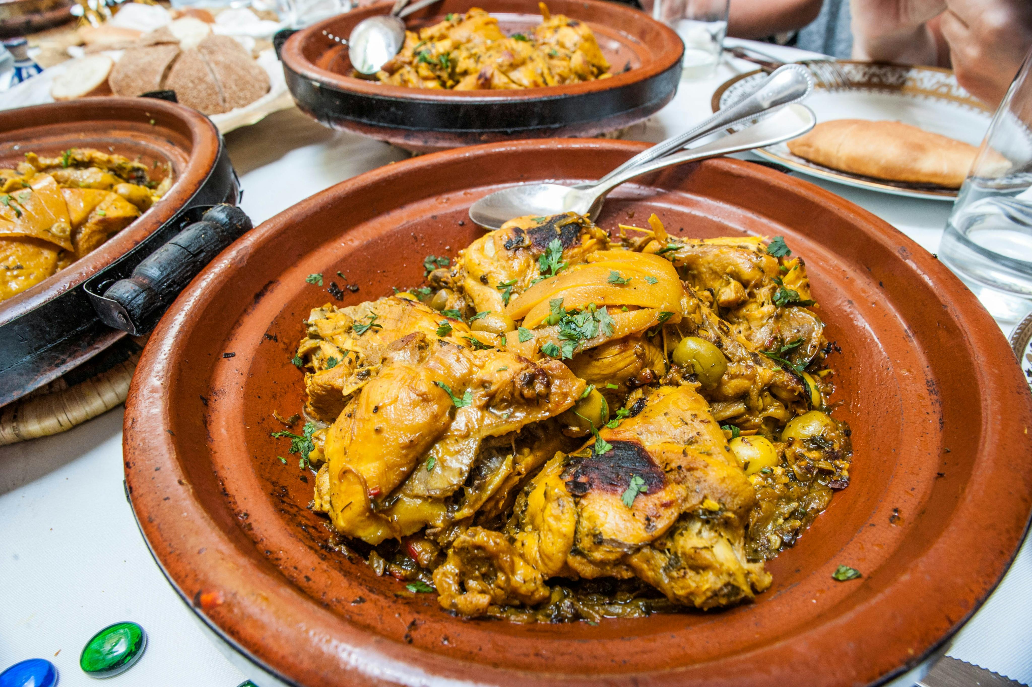 一道摩洛哥鸡肉塔吉恩，配有香草和香料，放在一个粘土锅里，里面放着一个勺子，桌子上放着额外的塔吉恩锅。