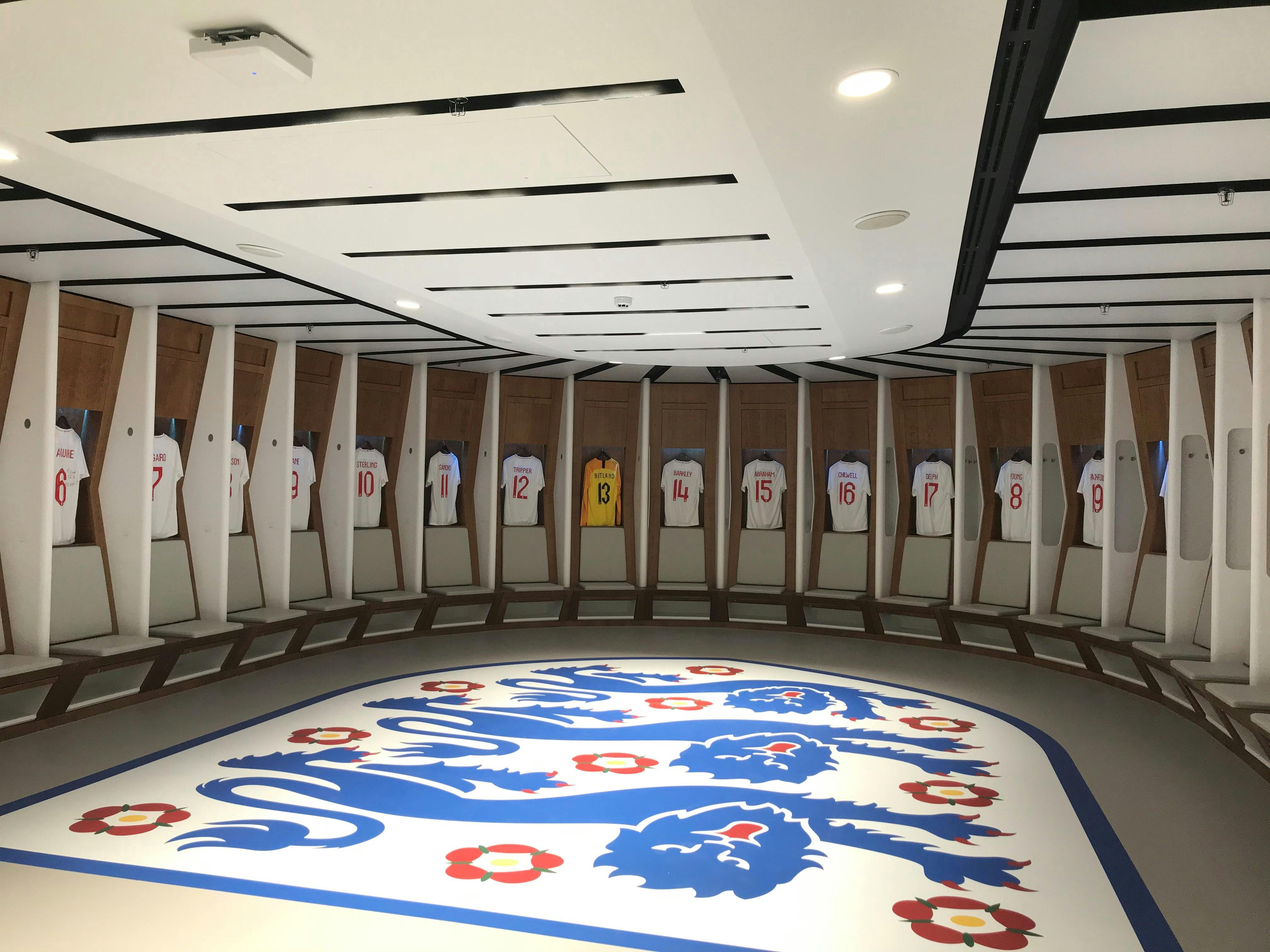 一间更衣室，座位上方悬挂着编号为1-20的白色足球衫，地板上挂着两只狮子的大型标志。
