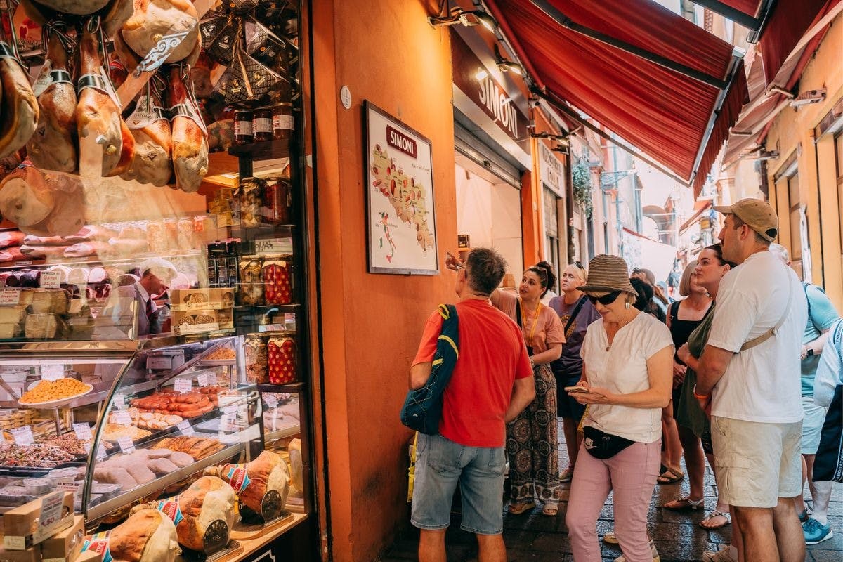 People in casual summer clothes gather outside a shop in a narrow alley with orange walls and red awnings.