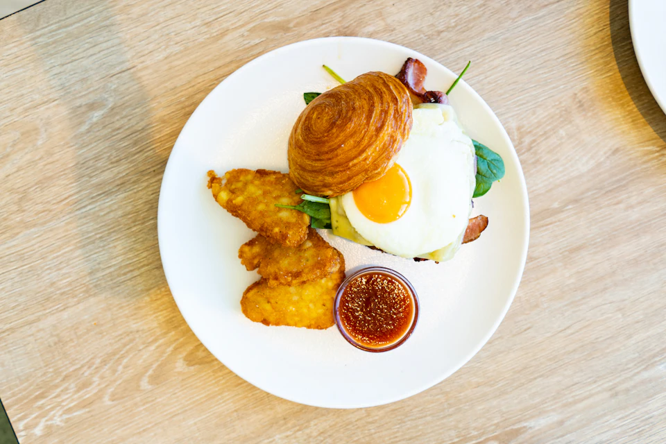 A breakfast plate with a croissant, sunny-side-up egg, bacon, hash browns, greens, and a small bowl of dipping sauce.