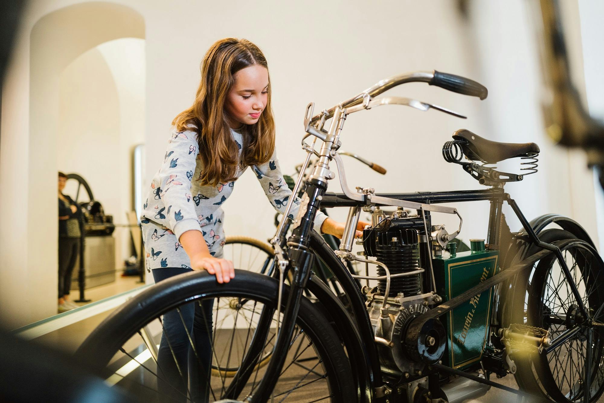 一个年轻的女孩在博物馆里检查一辆古董自行车，触摸它的车把，并倾向于它的发动机。