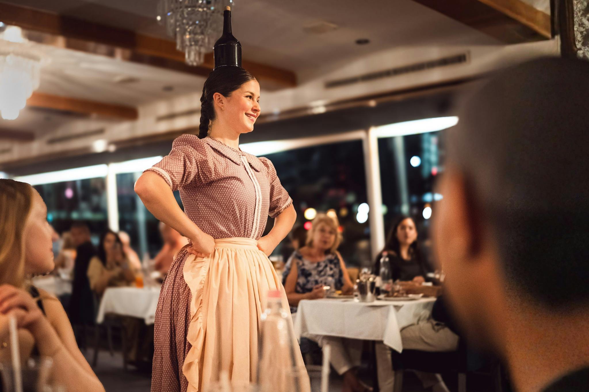 Eine Darstellerin in traditioneller Kleidung balanciert in einem Restaurant eine Flasche auf ihrem Kopf, während die Gäste von ihren Tischen aus zusehen.